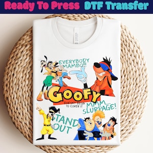 Op de afbeelding: Wit T-shirt met een kleurrijke Goofy cartoonafbeelding, inclusief het woord "Goofy" en zinnen als "Everybody Mambo!!!" en "Stand Out". Het shirt is op een geweven oppervlak gevouwen.