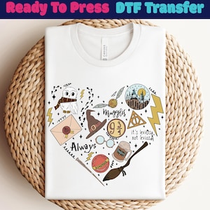 May include: White t-shirt with a heart-shaped design featuring illustrations of a wizarding world theme. The design includes an owl, lightning bolts, a hat, glasses, and the text "Always" and "Muggles". The shirt is on a woven surface.