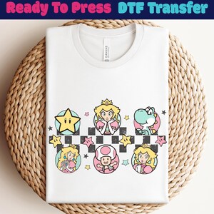 May include: White t-shirt with a colorful graphic featuring cartoon characters and the text "Ready To Press DTF Transfer". The design includes a star, Princess Peach, Yoshi, and a mushroom character, set against a checkered background.