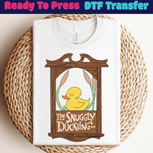 Könnte beinhalten: Weißes T-Shirt mit einer Grafik einer gelben Ente in einem Holzrahmen, mit dem Text "The Snuggly Duckling". Das Design beinhaltet Schilf und einen dekorativen Rahmen. Das T-Shirt ist auf einer gewebten Oberfläche gefaltet.