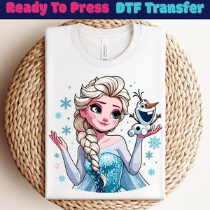 May include: White t-shirt with a cartoon image of Elsa and Olaf from Frozen, surrounded by snowflakes. The text "Ready To Press DTF Transfer" is at the top. The shirt is folded on a woven surface.