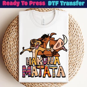 May include: White t-shirt featuring a colorful graphic of Timon and Pumbaa from The Lion King, with the words "Hakuna Matata" in a decorative font. The shirt is folded and placed on a woven basket.