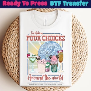 May include: White t-shirt with a colorful graphic design. The design includes the text "I'm Making POUR CHOICES Around the world" with illustrations of cocktails and a blue sphere. The shirt is folded on a woven mat.