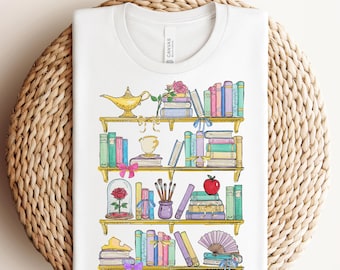 Puede incluir: Camiseta blanca con una colorida ilustración de una estantería llena de libros, una rosa y una lámpara. El texto "Ready To Press DTF Transfer" está en la parte superior. La camiseta está doblada sobre una cesta tejida.