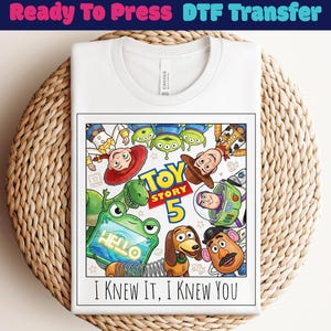 May include: A white t-shirt featuring a graphic print of Toy Story characters including Woody, Buzz Lightyear, Jessie, Rex, Slinky Dog, Mr. Potato Head, and aliens. The design includes the text Toy Story 5 and the phrase I KNEW IT, I KNEW YOU.