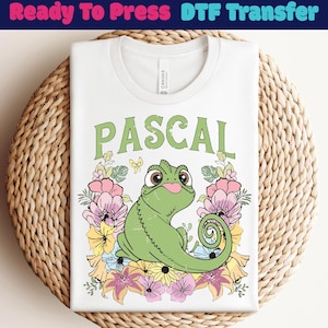 Könnte beinhalten: Weißes T-Shirt mit einem Aufdruck eines grünen Chamäleons namens Pascal, umgeben von bunten Blumen. Der Text "PASCAL" ist in grünen Buchstaben über dem Chamäleon aufgedruckt. Das T-Shirt ist auf einer runden, gewebten Unterlage gefaltet. Oben steht "Ready To Press DTF Transfer".
