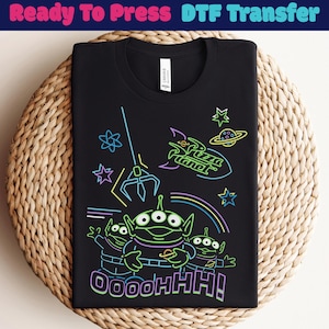 May include: Black t-shirt with a neon graphic of three aliens, a spaceship, and the words "Pizza Planet" and "Oooohhh!". The design includes stars and a claw. The shirt is displayed on a woven surface.