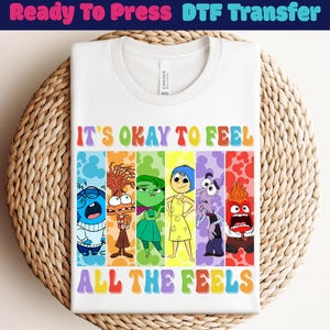 以下が含まれることがあります： カラフルな文字で「IT'S OKAY TO FEEL ALL THE FEELS」と書かれた白いTシャツ。さまざまな感情を表す漫画のキャラクターが描かれています。Tシャツは織られた表面に折りたたまれています。