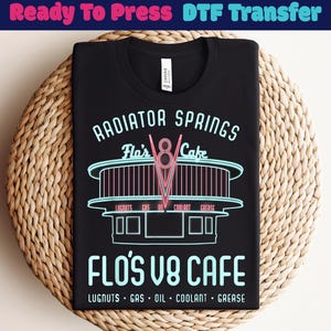 May include: Black t-shirt featuring a neon-style graphic of "Radiator Springs Flo's V8 Cafe". The design includes the cafe's name, a stylized "V8" logo, and the words "Lugnuts, Gas, Oil, Coolant, Grease".