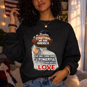 Puede incluir: Sudadera gris oscura con un diseño de balón de fútbol y el texto "TOGETHER, WE ARE AMERICA". Debajo, el texto dice "THE ONLY THING MORE POWERFUL THAN HATE IS LOVE". Se ve una bandera americana de fondo.