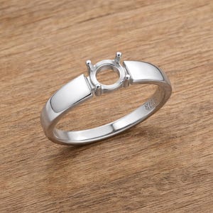 May include: A silver ring setting with a round center and four prongs. The ring has a polished finish and a simple band. The ring is set on a wooden surface.
