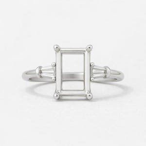 Puede incluir: Un engaste de anillo de compromiso plateado con una piedra central de talla esmeralda y dos piedras laterales baguette. El anillo presenta una abertura rectangular para la piedra central y pequeños detalles de cuentas. La banda es lisa y pulida.