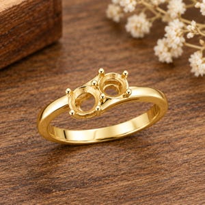 May include: A polished gold ring featuring a dual-setting design with two empty circular prongs arranged side-by-side. The band is smooth and rounded, showcasing a bright metallic finish throughout the entire structure of the jewelry piece.