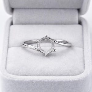 Puede incluir: Un engaste de anillo plateado con un centro redondo y seis pequeñas garras. El anillo se muestra en una caja de anillo de terciopelo gris. La banda se curva con gracia, creando un diseño moderno y elegante.