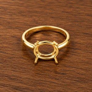 May include: A polished yellow gold ring setting featuring a smooth, rounded band and a four-prong oval head designed to hold a gemstone. The metal has a reflective, high-shine finish, showcasing a classic, clean structure for jewelry assembly.