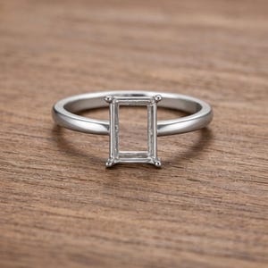 Puede incluir: Un engaste de anillo plateado con diseño de talla esmeralda. El anillo está sobre un fondo de madera. La banda del anillo es lisa y pulida, y el engaste está diseñado para sostener una gema rectangular.