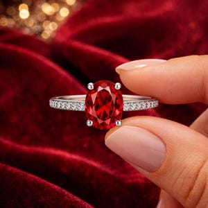 Può includere: Un anello d'argento con una grande gemma rossa brillante a taglio ovale come elemento centrale. La fascia è impreziosita da piccole pietre chiare e scintillanti. L'anello è presentato su uno sfondo di velluto rosso scuro.