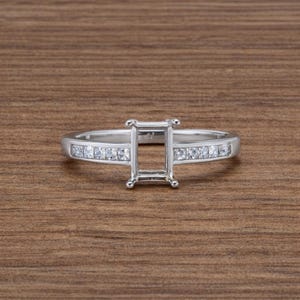 May include: A silver ring with a rectangular center setting and a band adorned with small, square-cut stones. The ring is set against a wooden background, showcasing its design and craftsmanship.