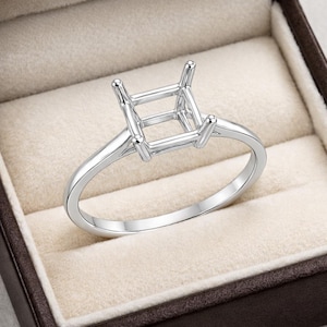 Puede incluir: Un anillo plateado con un engaste cuadrado para una piedra preciosa. El anillo tiene una banda delgada y cuatro garras en las esquinas del engaste. Se muestra en una caja de joyería beige.