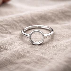 May include: A silver ring with a circular setting. The ring is set against a neutral-toned fabric background. The ring's design features a simple band and a round, open bezel, perfect for a custom gemstone or design.