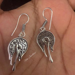 May include: A pair of silver-toned earrings featuring a melting clock design. Each earring has a hook, a coiled detail, and a circular face with clock numerals. The clock face appears to be dripping, creating a surreal effect.