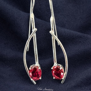 Könnte beinhalten: Ein Paar silberner Ohrhänger mit geschwungenem Design. Jeder Ohrhänger hat einen leuchtenden, oval geschliffenen, roten Edelstein. Die Ohrringe werden auf dunkelblauem Stoff präsentiert. Der Text "Vini Jewellers" ist sichtbar.