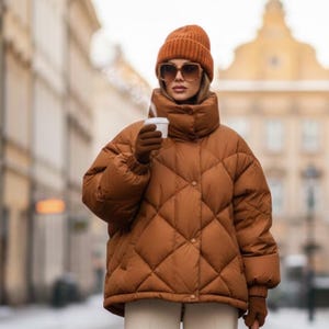 Könnte beinhalten: Eine Person trägt eine braune, gesteppte Pufferjacke, eine passende Strickmütze und Handschuhe und hält eine weiße Kaffeetasse. Die Jacke hat einen hohen Kragen und ein Rautenmuster. Die Sonnenbrille hat einen braunen Rahmen. Der Hintergrund zeigt eine Stadtstraße.