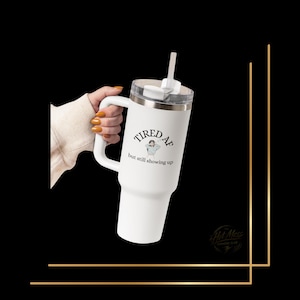 Puede incluir: Vaso térmico blanco con asa, borde plateado y pajita blanca. El vaso tiene el texto "TIRED AF but still showing up" con una pequeña ilustración de una persona.