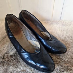 Può includere: Un paio di ballerine vintage in pelle nera con piccole fibbie decorative. Le scarpe hanno la punta arrotondata e un tacco basso. L'interno delle scarpe è di colore chiaro e sono appoggiate su una superficie strutturata simile alla pelliccia.