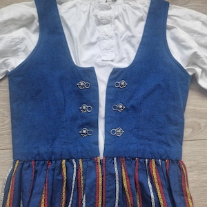 May include: A traditional blue vest with decorative silver clasps over a white blouse. The skirt features vertical stripes in red, yellow, and white. The outfit is a classic folk style.