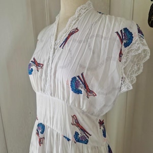Könnte beinhalten: Weißes Vintage-Kleid mit V-Ausschnitt, verziert mit einem sich wiederholenden Muster aus roten und blauen Figuren. Das Kleid hat eine Spitzenborte am Ausschnitt und an den Ärmeln sowie eine taillierte Taille. Der Stoff wirkt leicht und plissiert.