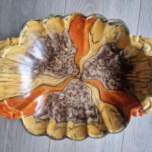 Könnte beinhalten: Dekorative Servierschale aus Keramik mit einer einzigartigen, organischen Form. Die Schale weist eine Mischung aus Farben auf, darunter Gelb-, Braun- und Orangetöne, mit Marmoreffekt. Sie hat Griffe und ist etwa 36 cm lang.