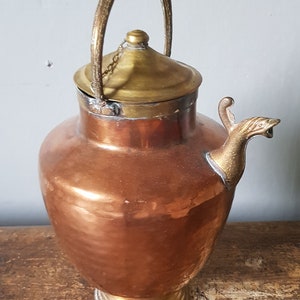 Könnte beinhalten: Eine Teekanne aus Kupfer und Messing mit einem dekorativen Ausguss und einem Henkel mit einer Kette. Die Teekanne steht auf einer Holzoberfläche.