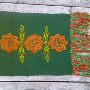 May include: A green wool throw blanket with orange and yellow floral embroidery and green and orange fringe.