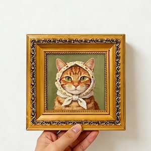 Könnte beinhalten: Ein Ölgemälde einer orangefarbenen getigerten Katze, die ein cremefarbenes Kopftuch mit kleinem Blumenmuster trägt. Die Katze hat grüne Augen und ist vor einem gedeckten grünen Hintergrund zentriert. Das Bild ist in einem verzierten Goldrahmen.