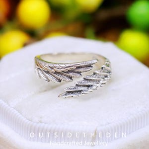 May include: A silver-colored ring with a wing design. The ring features detailed feather textures, with the wing wrapping around the finger. The ring is displayed on a white surface. The text "OUTSIDETHEDEN Handcrafted Jewellery" is visible.