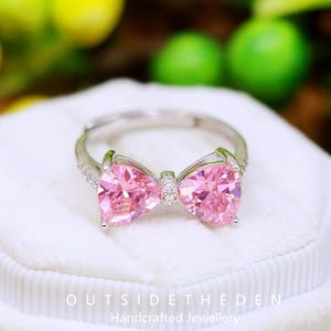 Può includere: Anello color argento con un design a forma di fiocco con due grandi pietre rosa a forma di pera. L'anello ha una fascia delicata ed è posto su una superficie bianca. Il testo "OUTSIDETHEDEN Handcrafted Jewellery" è visibile in basso.