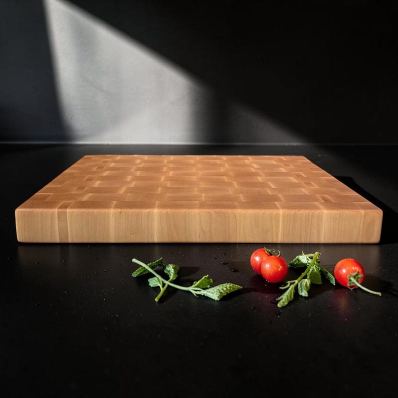 May include: A rectangular end-grain wooden cutting board with a light tan color and a visible grid pattern of wood blocks. Two small red cherry tomatoes with green stems are positioned in front of the board on a dark surface.