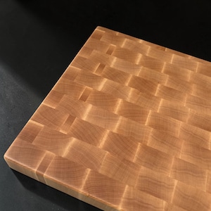 May include: A rectangular wooden cutting board featuring an end-grain construction with a woven pattern of light brown wood blocks. The surface displays natural wood grain variations and a smooth finish across the interlocking square segments.