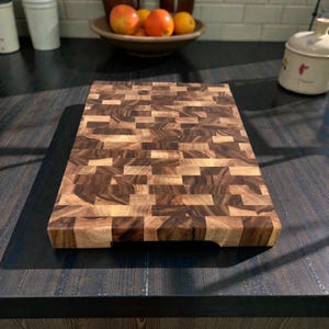 Puede incluir: Una tabla de cortar rectangular de grano final con un patrón de mosaico de bloques de madera claros y oscuros. La tabla tiene una superficie lisa y bordes redondeados, con un asa recortada en un lado. Los tonos de madera van del beige claro al marrón oscuro.