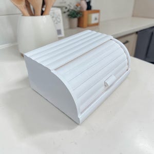 May include: A white wooden bread bin with a roll-top lid. The bin has a rectangular base and curved sides. The lid features horizontal, grooved slats. A small, rectangular handle is on the front. The bread bin is on a white countertop.