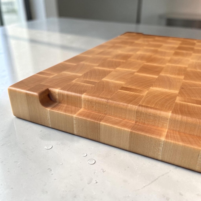 Close-up of the maple end grain board's corner on marble, showing the carved finger-grip handle groove and the thick edge in detail.
