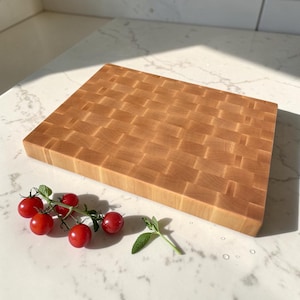 Maple end grain butcher block angled on a sunlit marble counter, with cherry tomatoes on the vine and a rosemary sprig arranged in front.