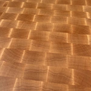 May include: A light brown wooden cutting board featuring an end-grain construction with a repeating rectangular block pattern. The surface displays a natural wood grain with varying light and dark tones, creating a textured, grid-like appearance.