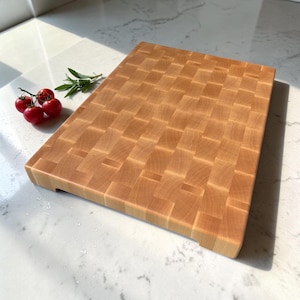 Angled view of the thick maple end grain chopping block on a marble counter, its deep edge in focus, with cherry tomatoes and rosemary to the side.