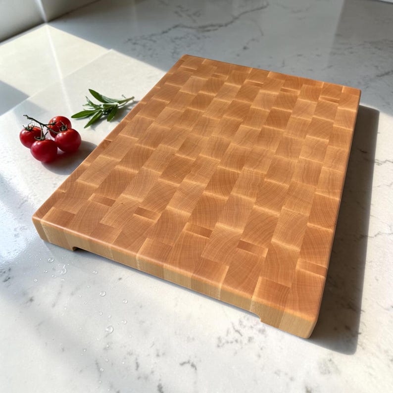 Angled view of the thick maple end grain chopping block on a marble counter, its deep edge in focus, with cherry tomatoes and rosemary to the side.