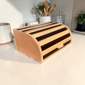 Op de afbeelding: Houten brooddoos met een gebogen bovenkant en een lichtbruin en donkerbruin gestreept ontwerp. De doos heeft een klein houten handvat aan de voorkant en is gemaakt van natuurlijk hout.