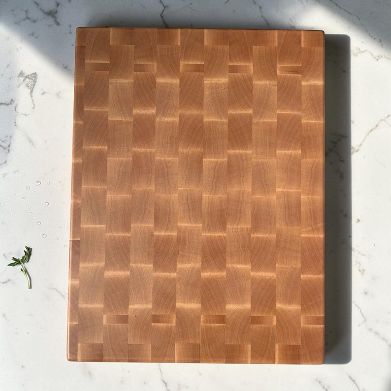 Overhead close-up of the maple end grain board filling the frame on marble, highlighting the fine checkerboard grain, with a small basil sprig at the corner.