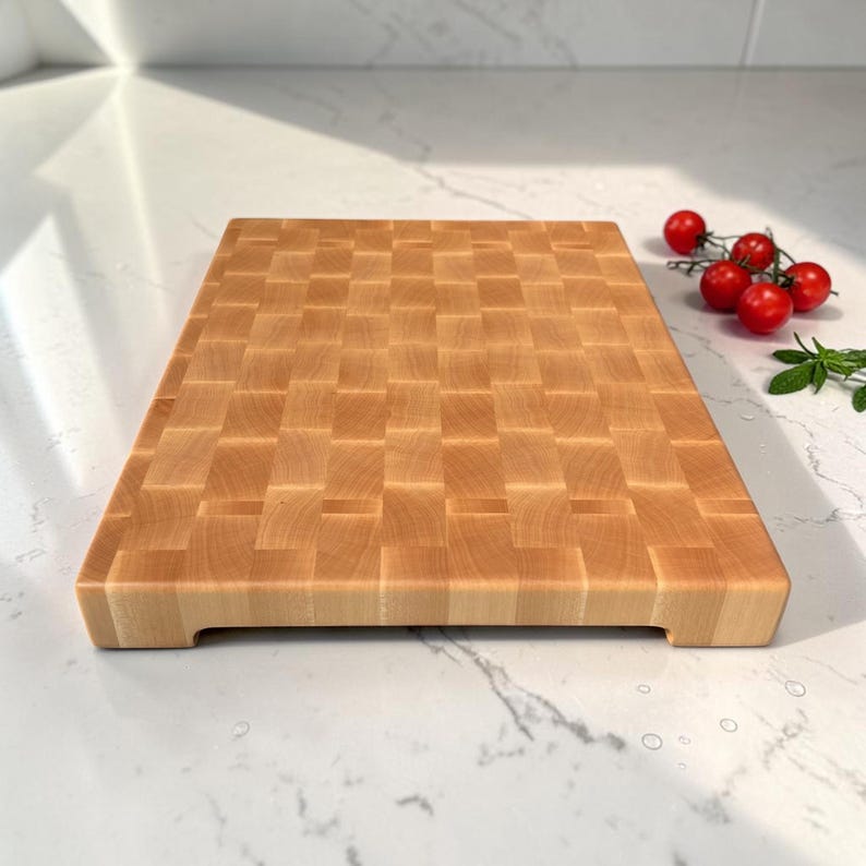 Maple end grain cutting board lying flat on a sunlit marble counter, top view showing the full checkerboard surface and side handle grooves, with cherry tomatoes at the corner.