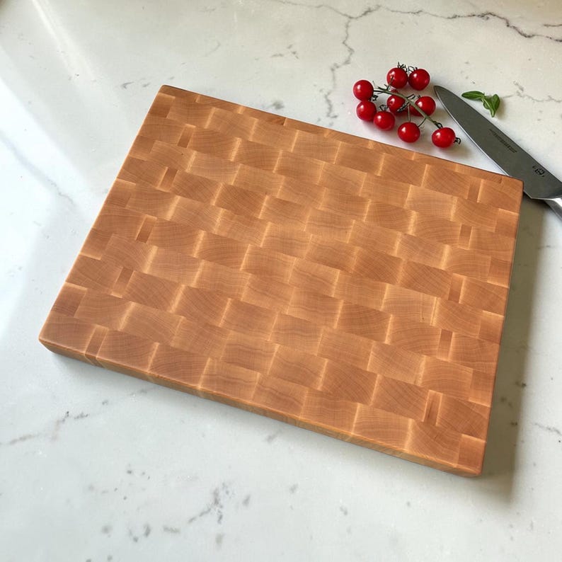 Large end grain maple cutting board on a white marble countertop, with cherry tomatoes on the vine and a kitchen knife beside it.